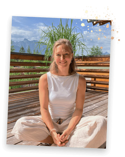Johanna sitzt lächelnd im Schneidersitz auf einer Terrasse mit Bergblick.